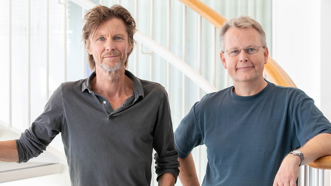 Two men standing indoors near a staircase, smiling at the camera.